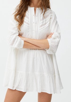 Femme portant une robe blanche à manches longues avec des détails en dentelle et un ourlet à volants, bras croisés devant son torse.