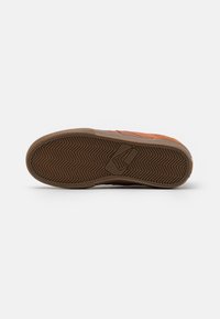 Semelle de chaussure présentant un motif en chevrons en caoutchouc marron, avec une section lisse pour l'adhérence ; accentuée par un revêtement orange sur le côté.