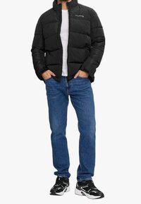 Chaqueta negra acolchada con cuello alto, logo blanco, llevada sobre una camisa blanca, combinada con jeans azules y zapatillas deportivas negras.