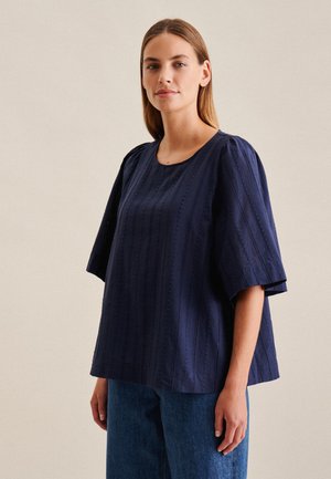 Top in cotone blu navy con scollatura rotonda, maniche corte a sbuffo e strisce verticali testurizzate. Abbinato a pantaloni di jeans azzurri.