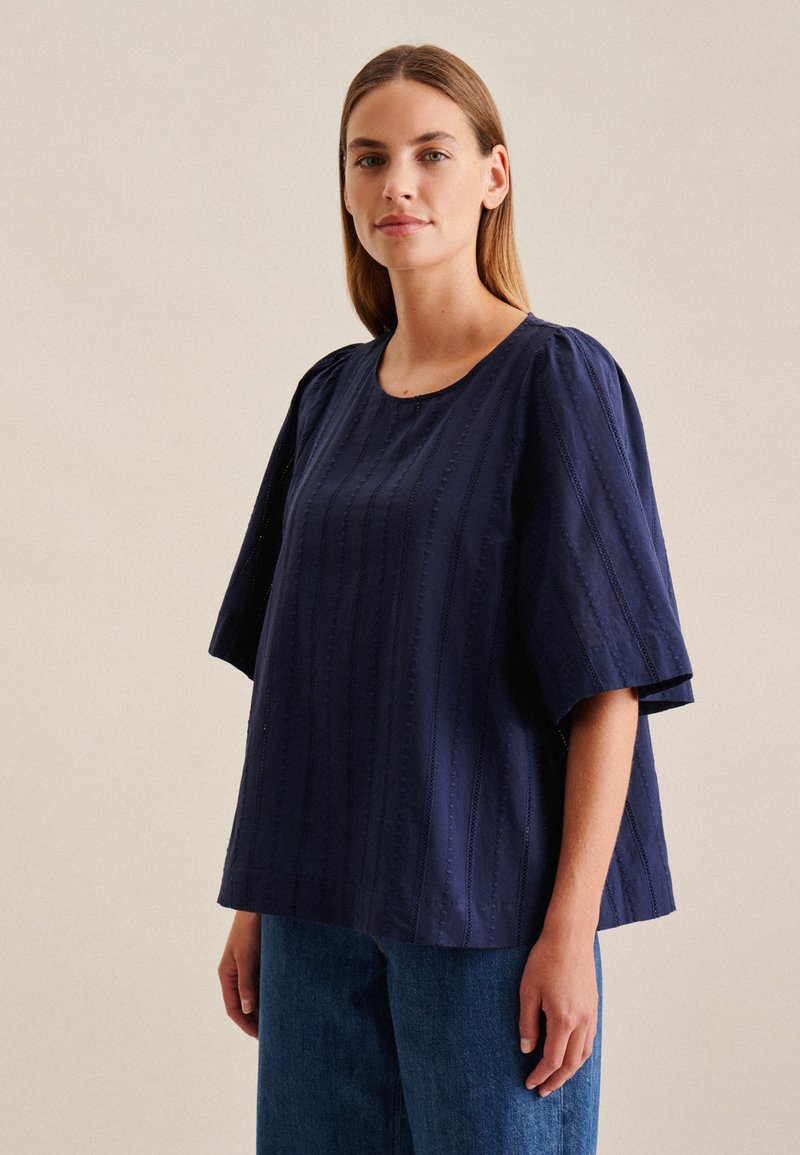Top in cotone blu navy con scollatura rotonda, maniche corte a sbuffo e strisce verticali testurizzate. Abbinato a pantaloni di jeans azzurri.