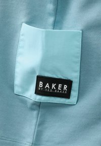 Světle modrá látka s texturovaným povrchem, na kterém je hladká kapsa s černou etiketou se nápisem "BAKER by TED BAKER."