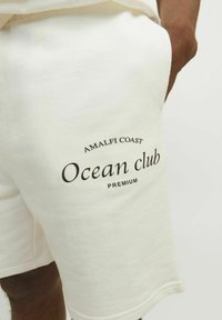 Bela bomba iz bombaža s mehakim teksturo, z natisnjenim črnim besedilom: "AMALFI COAST Ocean club PREMIUM." Preprost dizajn, dolžina do kolen.