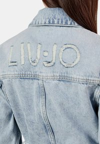 Giacca in denim azzurro chiaro con dettagli sfilacciati e ricamo "LIV·JO" sulla schiena, dotata di colletto classico e cuciture a vista.