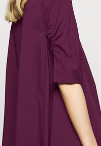 Burgundy klänning med samlade ärmar, som har en avslappnad passform och slät textur. Korta ärmar med rullad ärmdetalj.