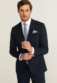 Costume bleu marine à la coupe ajustée, avec une texture lisse, chemise blanche, cravate bleu clair et pochette à motifs.