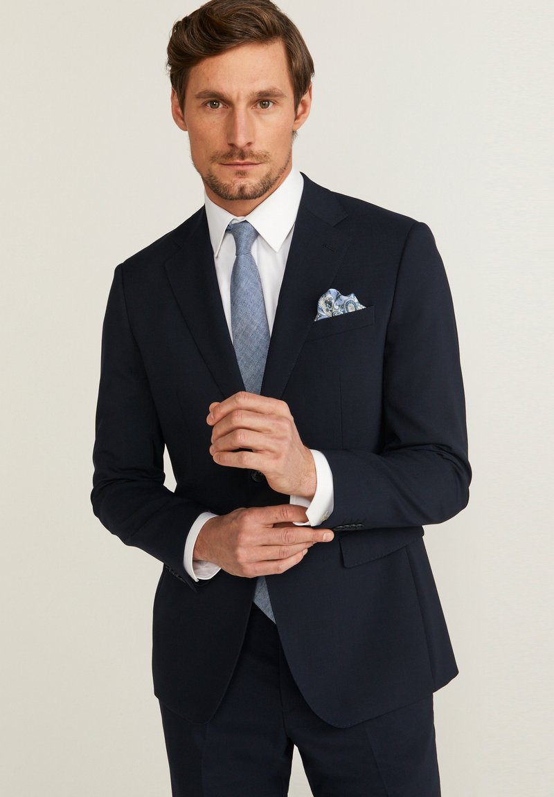 Costume bleu marine à la coupe ajustée, avec une texture lisse, chemise blanche, cravate bleu clair et pochette à motifs.