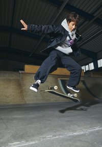 Skateboardåka i svart jacka och grå hoodie hoppar upp i luften över en skateboardramp. Svarta byxor och skor, träskateboard med ljusa hjul.