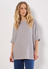 Jasnoszara, oversize'owa dzianinowa bluzka z teksturowym wzorem, kr�ótkimi, podwiniętymi rękawami i szerokim dekoltem, zestawiona z niebieskimi dżinsami.