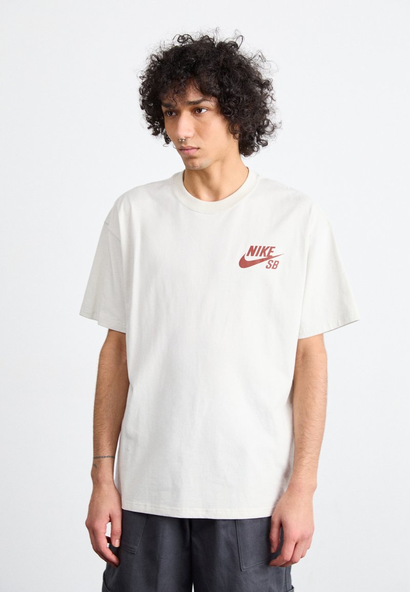 Krémeové bavlnené tričko s krátkymi rukávmi, okrúhlym výstrihom a červeným logom Nike SB na ľavej hrudi. Hladká textúra, uvoľnený strih.