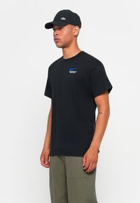 Fekete pamut póló rövid ujjakkal és kerek nyakkivágással, kék Nike logóval és "Just Do It" felirattal. Olíva színű sapkával párosítva.