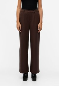 Pantalon large marron à texture lisse, avec taille haute, plis à l'avant et poches latérales ; porté avec des chaussures à plateforme noires.