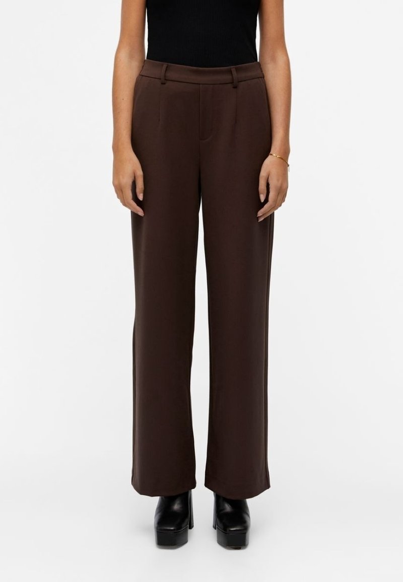 Pantalon large marron à texture lisse, avec taille haute, plis à l'avant et poches latérales ; porté avec des chaussures à plateforme noires.
