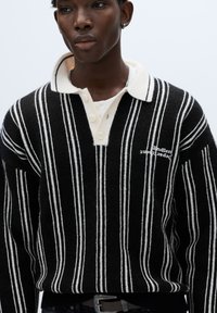 Maglione a righe bianche e nere con scollo a collo, dotato di una patta con bottoni bianchi e dettaglio di testo ricamato sulla parte anteriore.