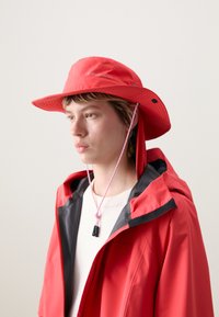 Chapeau de seau imperméable rouge avec un large bord, cordon ajustable rose et rabats pour les oreilles, associé à une veste rouge assortie avec des accents noirs.