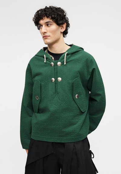 Νεαρός άνδρας με σγουρά σκούρα μαλλιά, φορώντας πράσινο υφασμάτινο φούτερ με μεταλλικά κουμπιά και μεγάλες τσέπες με καπάκια, κοιτάζοντας προς τα πλάγια.