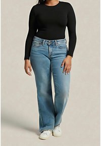 Femme portant un haut noir ajusté à manches longues, un jean taille haute bleu clair et des baskets blanches, debout devant un fond uni.
