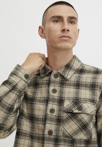Chemise en flanelle à carreaux beige et marron avec boutons et poche poitrine. Tissu texturé, col pointu et détails de coutures visibles.