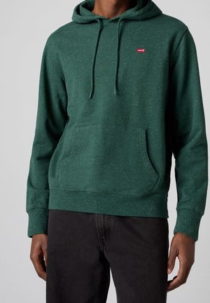 Mann trägt einen dunkelgrünen gesprenkelten Hoodie mit Fronttasche und Levi’s-Logo auf der Brust, kombiniert mit schwarzen Hosen, vor einem einfarbigen Hintergrund.