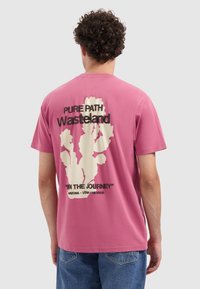 Kurzes pinkes Baumwoll-T-Shirt mit einem weißen Kaktus-Design, schwarzem Text "Pure Path Wasteland" und "In The Journey" auf der Rückseite.
