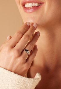 Zilveren ring met twee in elkaar grijpen banden, met een gladde afwerking en textuuraccenten. Hand die zachtjes de kin aanraakt; zacht stof op de achtergrond.