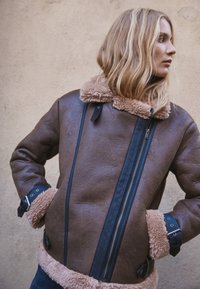 Bruine shearling jas met zwarte accenten, voorzien van een bontkraag, ritssluiting en banddetails bij de manchetten. Gehele jas met een gestructureerde afwerking.