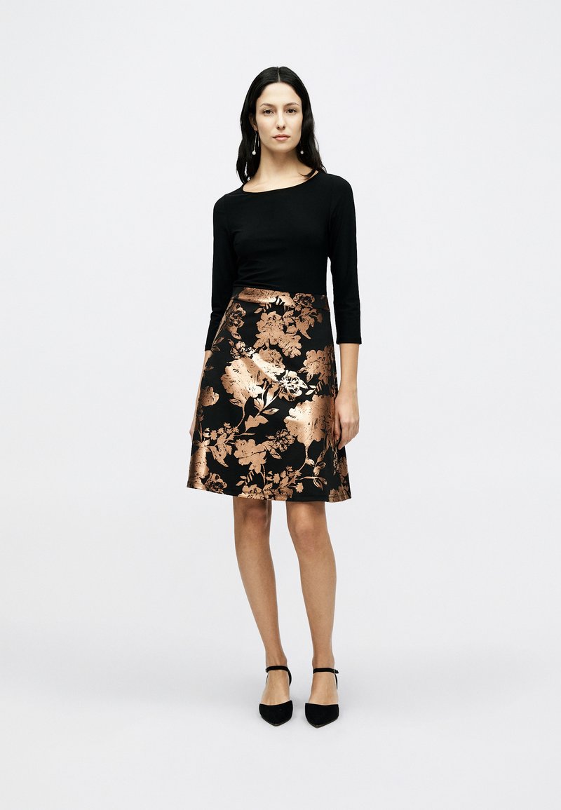 Μαύρο μακρυμάνικο τοπ σε συνδυασμό με φούστα μήκους μέχρι το γόνατο, διακοσμημένη με μεταλλικά ροζ χρυσά λουλουδάτα μοτίβα και λεία υφή. Φορεμένο με μαύρα τακούνια.