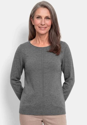 Vrouw met lang grijs haar, kleding een grijze gebreide trui en beige broek, staat met haar handen achter haar rug en glimlacht zachtjes.