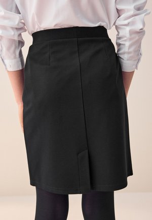 Czarna spódnica do kolan z ukrytym zamkiem błyskawicznym, z rozporkiem z tyłu i gładką teksturą materiału. Połączona z ciemnymi rajstopami.