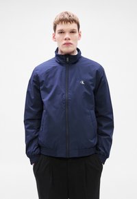 Μπλε ναυτικό μπουφάν με φερμουάρ, υψηλό γιακά, ελαστικές μανσέτες και μέση. Φέρει διακριτικό λογότυπο CK στο στήθος. Ομαλό, ελαφρύ ύφασμα.