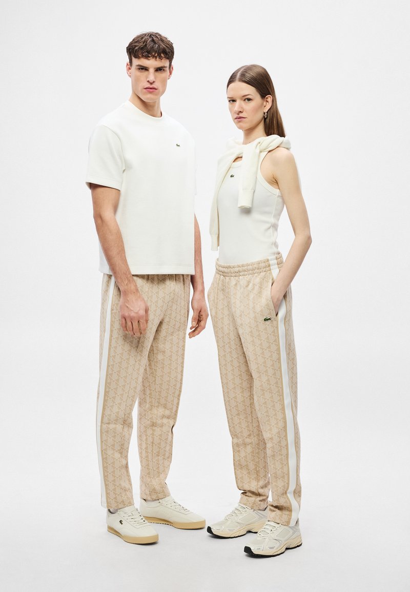 Maglietta bianca a maniche corte, pantaloni beige con motivi a strisce bianche e scarpe da ginnastica chiare. Design semplice con un logo discreto sia sulla maglietta che sui pantaloni.