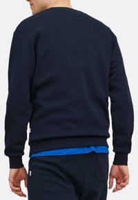 Sweatshirt bleu marine avec un col rond, des poignets et un ourlet côtelés. Porté sur une chemise bleue, fabriqué dans un tissu doux et texturé. Design simple et minimal.
