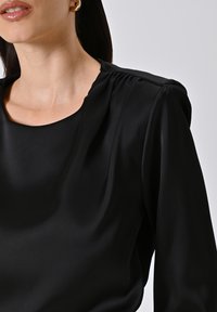 Blusa in satin nera con scollatura rotonda, morbidi drappeggi sulle spalle e texture liscia. Presenta un design aderente con maniche lunghe.