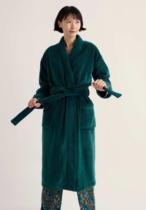 Femme portant une robe de chambre vert foncé et un pantalon à motifs, tenant et ajustant la ceinture de la robe sur un fond uni.