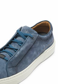 Blå mockaskor med vit gummisåle, med blå snören och en subtil logotyp på sidan. Slät innersula synlig.