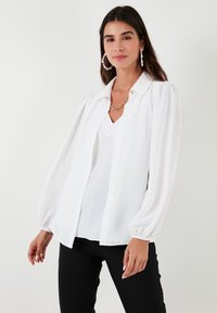 White blouse with a collar, long puffed sleeves, and a button front. Worn over a V-neck top, paired with black pants.