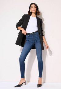 Blazer negro con mangas remangadas sobre una camiseta blanca, combinado con jeans ajustados de talle alto en azul y tacones negros de punta.