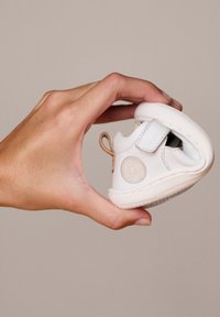 Chaussure pour bébé en cuir blanc tenue dans une main, avec une sangle Velcro, un logo circulaire, une forme arrondie et une semelle en caoutchouc texturée.