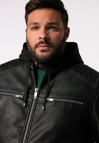 Giacca in pelle nera con cappuccio, dotata di tasche con zip, accenti a coste sulle spalle e finitura testurizzata. Modello che indossa una maglietta verde sotto.