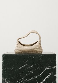 Woven beżowa torebka z płaskim dnem i zaokrąglonym uchwytem, o teksturowanym wzorze. Umieszczona na czarnym marmurowym postumencie.