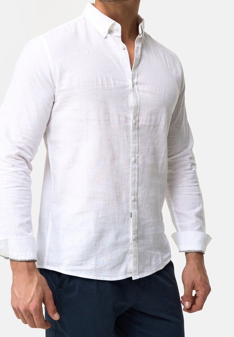 Homme portant une chemise blanche cintrée à manches longues avec un pantalon sombre, debout devant un fond clair uni.