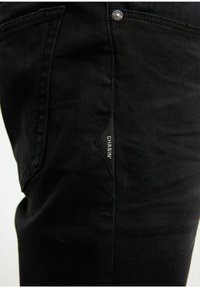 Pantalones de mezclilla negra con corte ajustado, que cuentan con un pequeño logo blanco en el lateral y un botón plateado en la cinturilla. Textura suave y diseño minimalista.