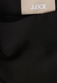 Černá tkanina s hladkým povrchem a jemným leskem, opatřená béžovou nálepkou s textem "JJXX" v bílé barvě.