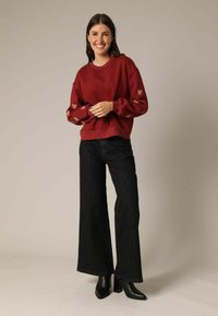 Sweat-shirt rouge avec des accents en forme de cœur sur les manches, associé à un jean noir à jambes larges. Modèle portant des bottines noires à talons. Fonds neutres.