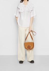 Blouse blanche avec col à volants et manches courtes, associée à un pantalon large crème. Sac marron avec un accent circulaire et une sangle réglable.