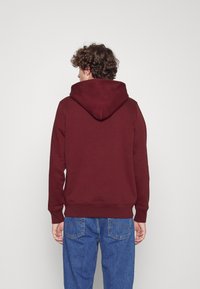 Felpa cappuccio bordeaux con tasca a marsupio e polsini a costine, abbinata a jeans di denim blu. Il tessuto appare morbido con una texture liscia.