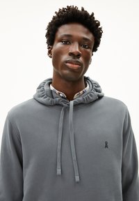 Grijze hoodie met een trekkoordkap, gemaakt van een textuurstof en met een klein geborduurd logo op de borst. Onderstaande col shirt is zichtbaar.