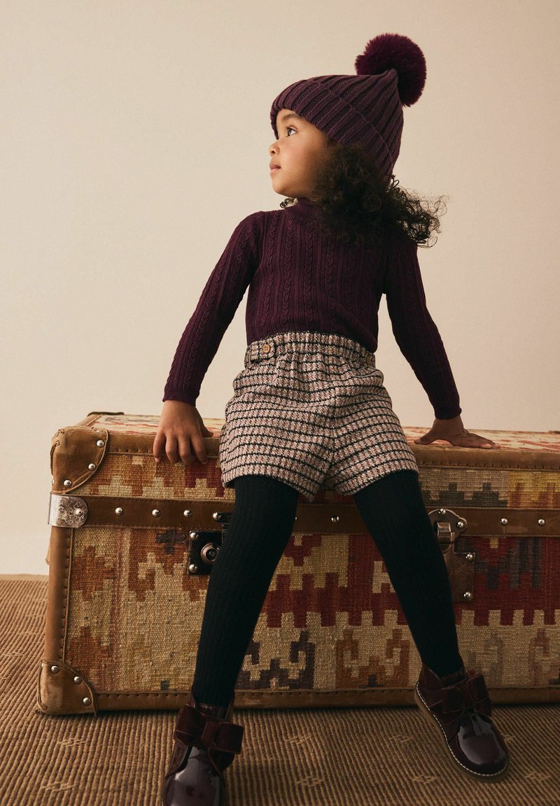 Jeune enfant portant un chapeau tricoté violet et un pull, un short à carreaux, des collants noirs et des bottes, assis sur un coffre vintage à motifs.