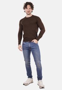 Uomo in piedi con una mano in tasca, indossando una camicia marrone a maniche lunghe, jeans blu e sneaker bianche su uno sfondo bianco puro.