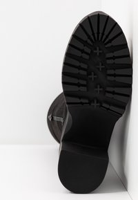 Semelle de botte noire avec des crampons profonds et des motifs en forme de croix, photographiée en position verticale sur un fond blanc.